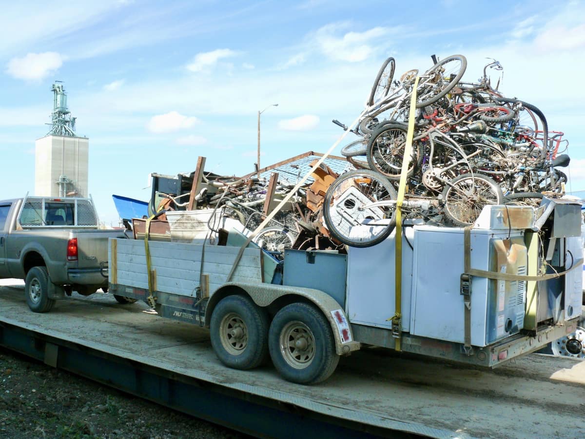 A large pile of scrap bicycles on a flat deck trailer. The bicycles are stacked on top of broken laundry machines. The is a bunch of other scrap metal piled up on the trailer and everything is strapped down.